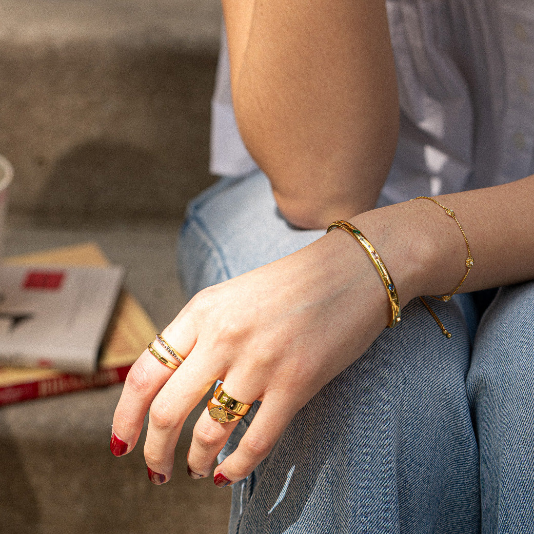 A person with red nail polish wears gold rings, the Hidden Curse Word Fuckin A Bracelet (a Local Eclectic exclusive), and a bangle on their hand resting on blue jeans; a book and takeaway coffee cup are in the background on concrete steps.