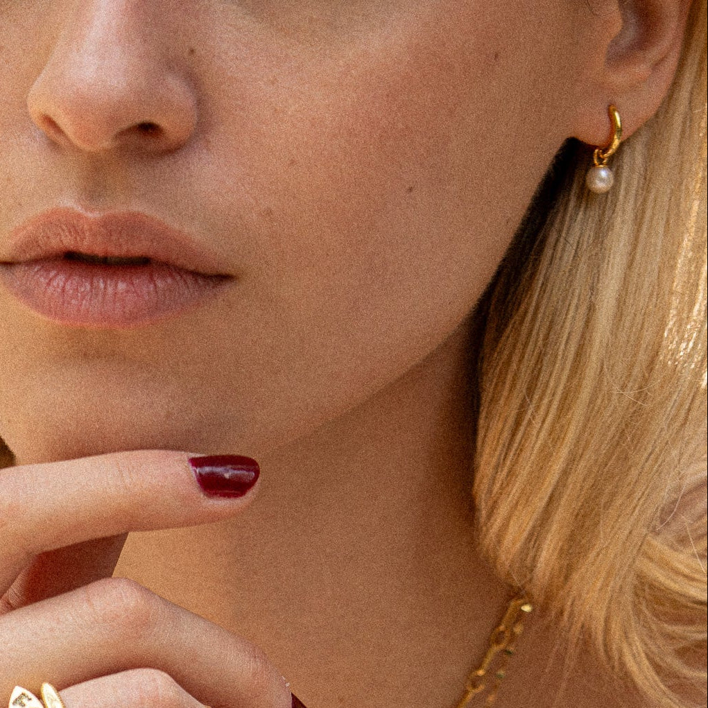 A close-up of a fair-skinned woman’s lower face, lips, and chin shows her blonde hair, maroon nail polish, gold ring, and Pearl Keepsake Huggie Hoops earrings. She also wears a 14k gold vermeil necklace.