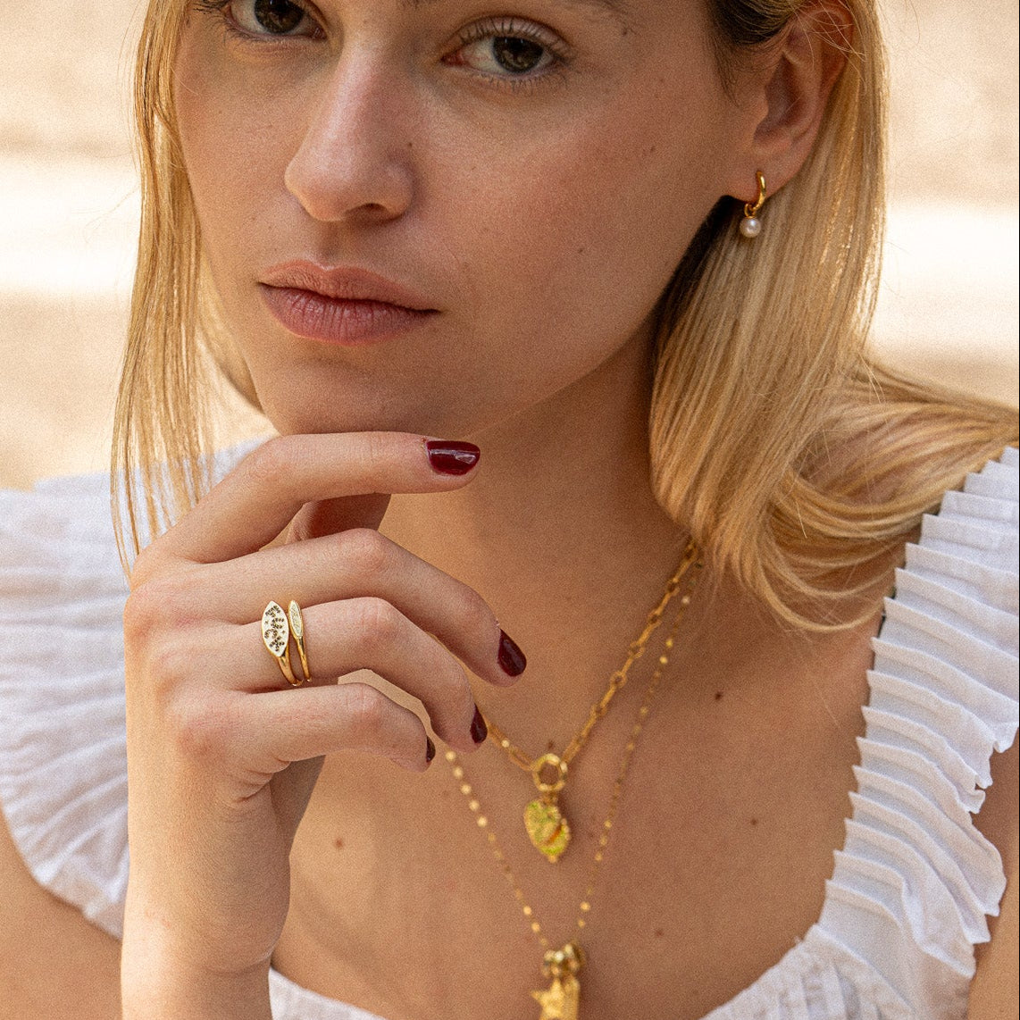 A blonde woman in a white ruffled top wears the Colorful Language Fuck Signet Ring and 14k yellow gold plated jewelry, including two necklaces and an earring, resting her hand near her face as she gazes thoughtfully at the camera.