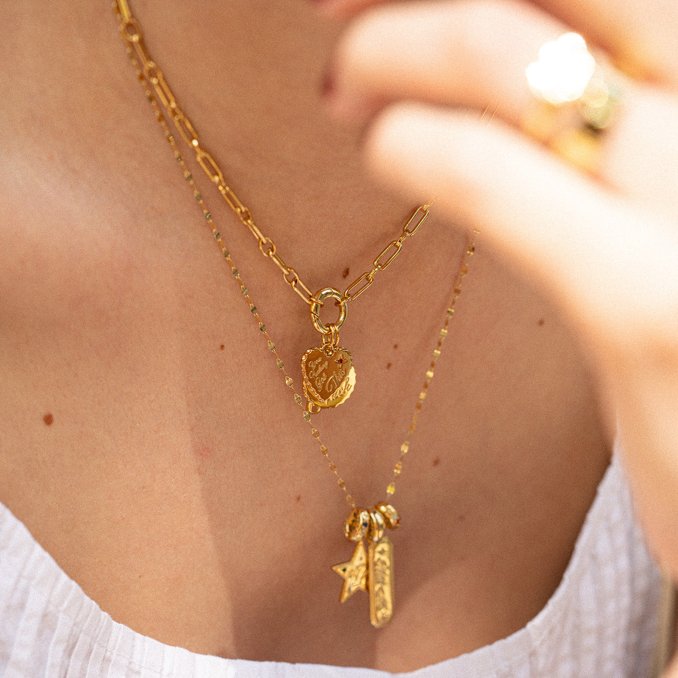 Close-up of a person wearing two layered necklaces, crafted in 14k yellow gold plating—one with a heart locket, the other featuring the Curse Word Charm plus a star and rectangular pendants, styled over a white textured top.