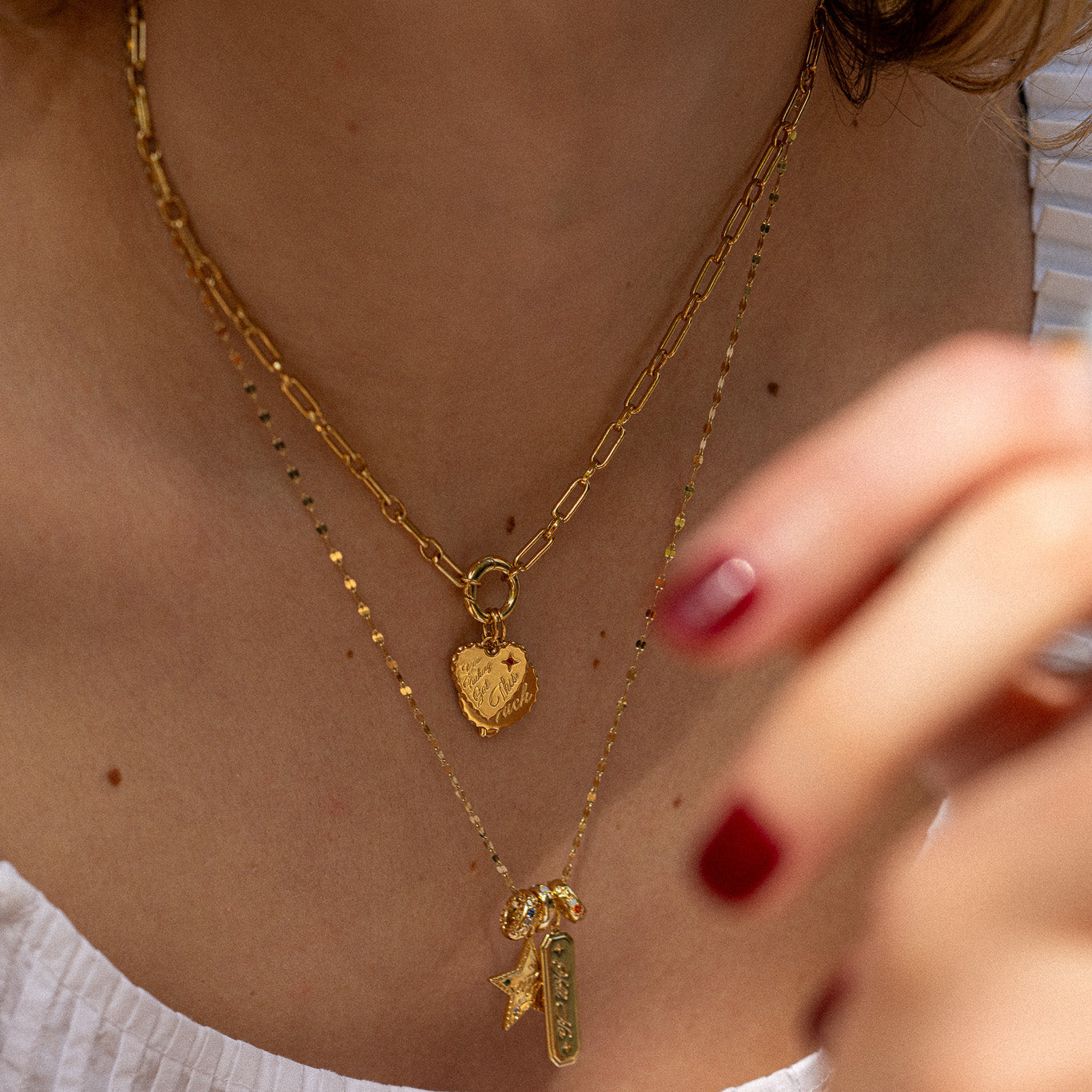 A woman with wavy hair, red nails, and a white top wears layered necklaces including the Curse Word Charm and a 14k yellow gold plated heart-shaped locket. Another necklace features a star and small pendant. Close-up shot.