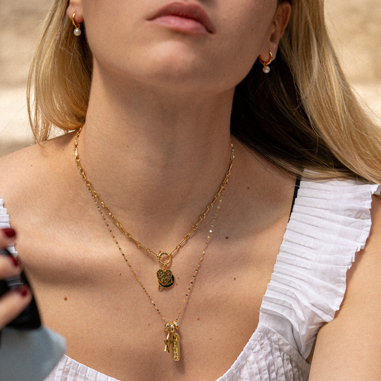A woman with long blonde hair wears gold hoop earrings and a gold-plated necklace featuring the Hidden Curse Word Spacer Charm. She’s dressed in a white ruffled sleeveless top, holding a phone, with her face partially out of frame.