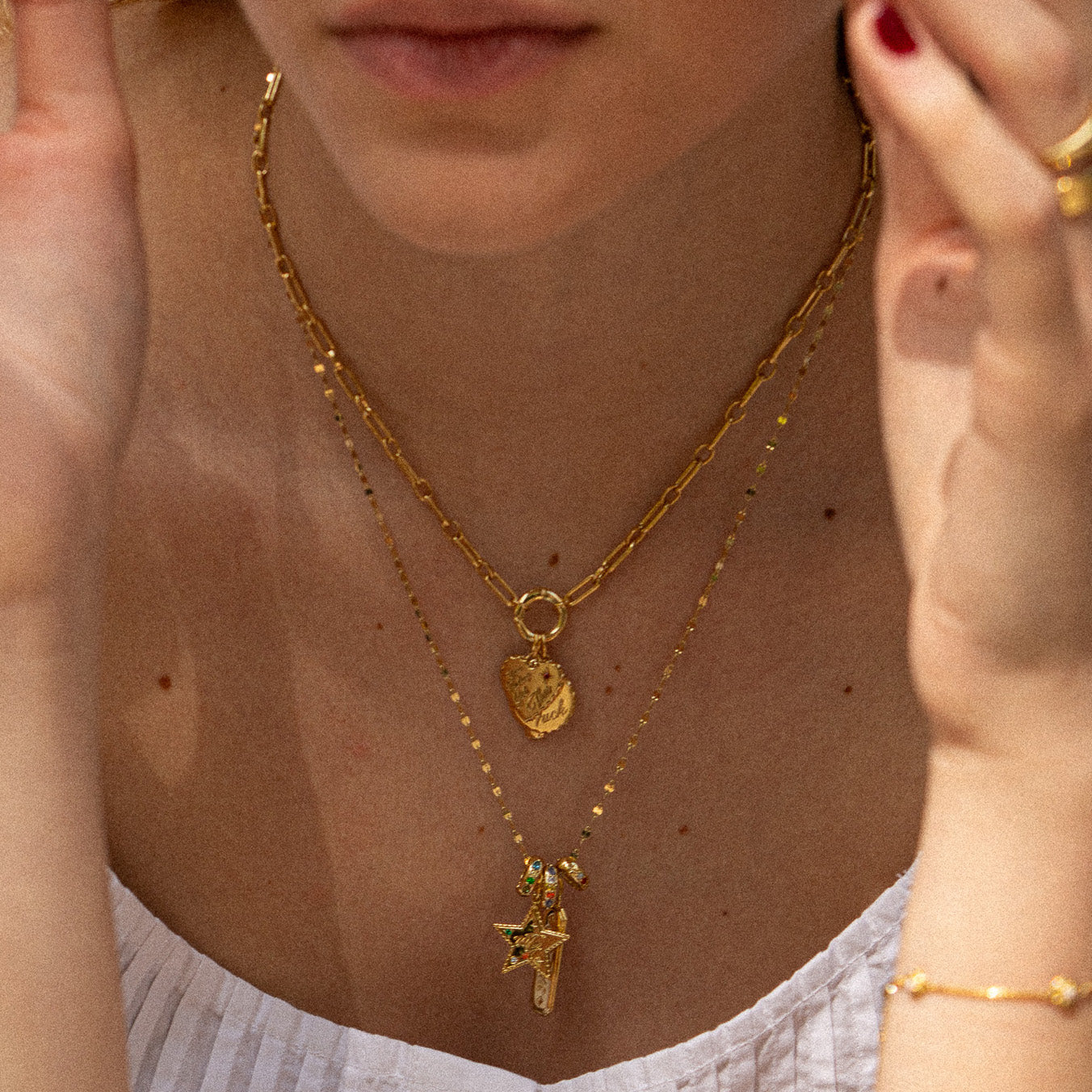A woman with short hair, red nails, and painted lips wears a white sleeveless top and two layered gold-plated necklaces—one featuring the Hidden Curse Word Spacer Charm and another with a round pendant.