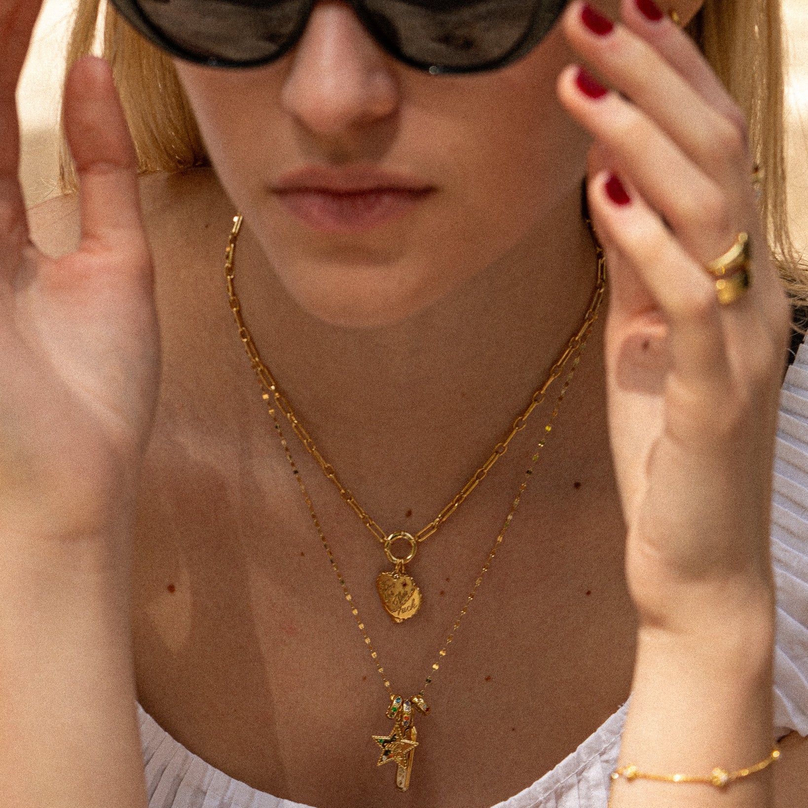 A woman in sunglasses and a white top adjusts her hair, highlighting red nail polish, layered gold necklaces—including the Curse Word Charm—and a gold bracelet. The focus is on her jewelry and hands.
