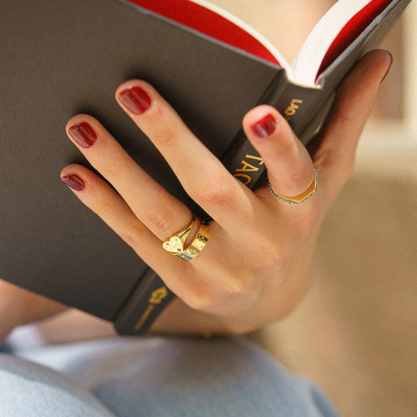 A hand adorned with red nail polish and the bold Tiny Cunt Signet Ring, a 14k gold plated heart-shaped statement piece, holds an open black book. The rings and manicured hand are in focus against a softly blurred, elegant background.