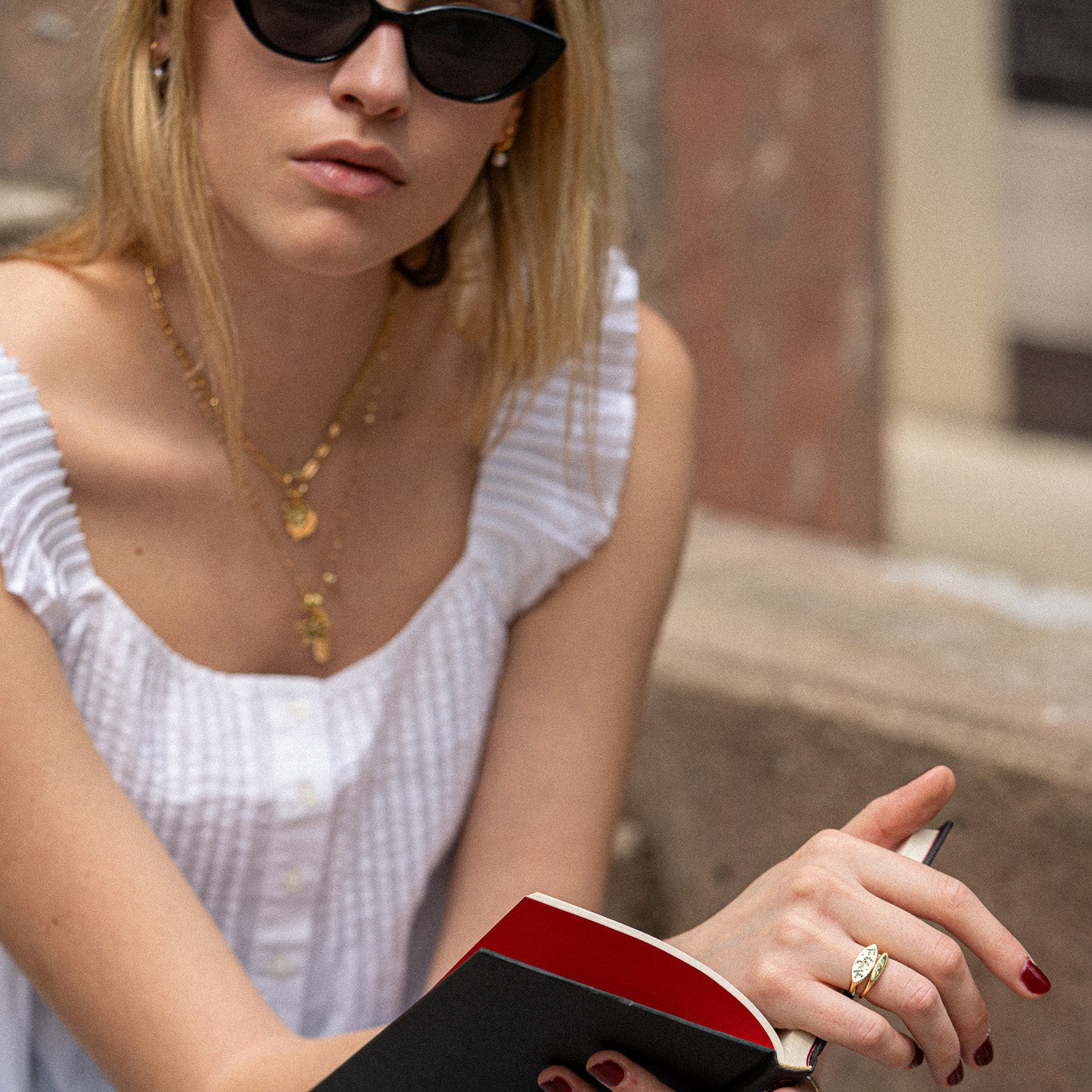 A relaxed woman in sunglasses and a white top sits outdoors reading, her blond hair loose. She wears gold jewelry, including the Colorful Language Fuck Signet Ring and a necklace, along with red nail polish.