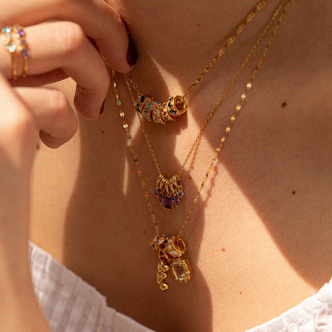 Close-up of a person in a white textured top wearing layered gold necklaces, including the Shades of You Ombre Birthstone Charm Necklace with colorful pendants and an ombre birthstone ring.