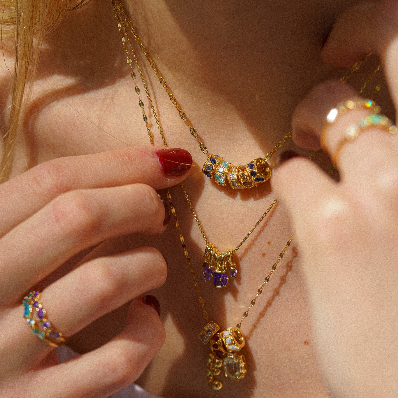 Close-up of a person wearing layered gold necklaces and gemstone rings, touching the necklaces while showing off red-painted nails. The focal piece is the Shades of You Birthstone Spacer Charm with colorful gemstones.