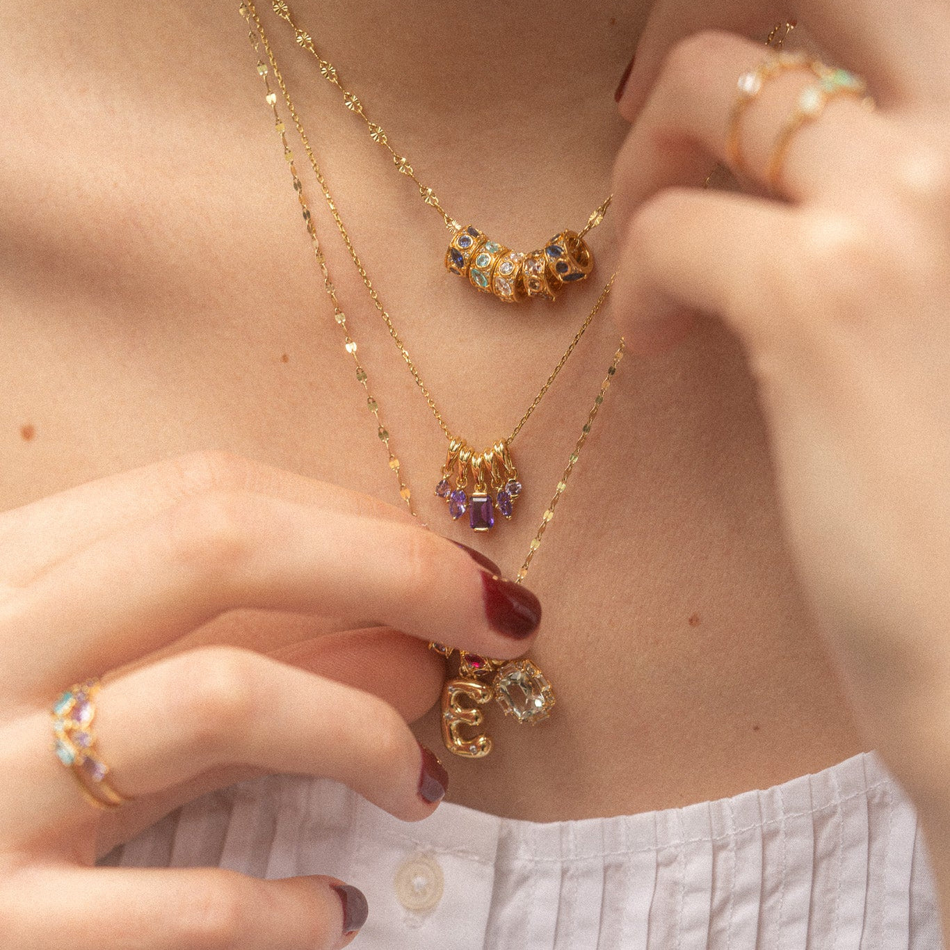 Wearing layered gold necklaces with colorful beaded charms, the Star Crossed Celestial Bubble Letter Charm, and initial pendants, plus gemstone-adorned gold rings, someone adjusts their jewelry over a white textured top.