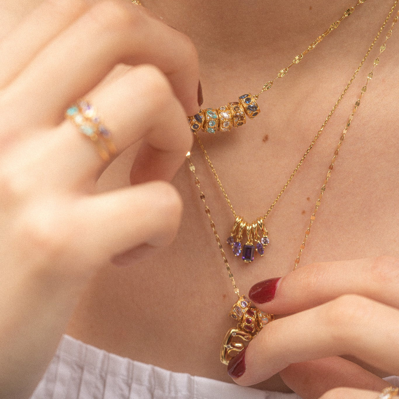 Close-up of a person wearing layered gold necklaces featuring the Shades of You Birthstone Spacer Charm and a matching gold ring. Their nails are painted red and they are dressed in a white top.