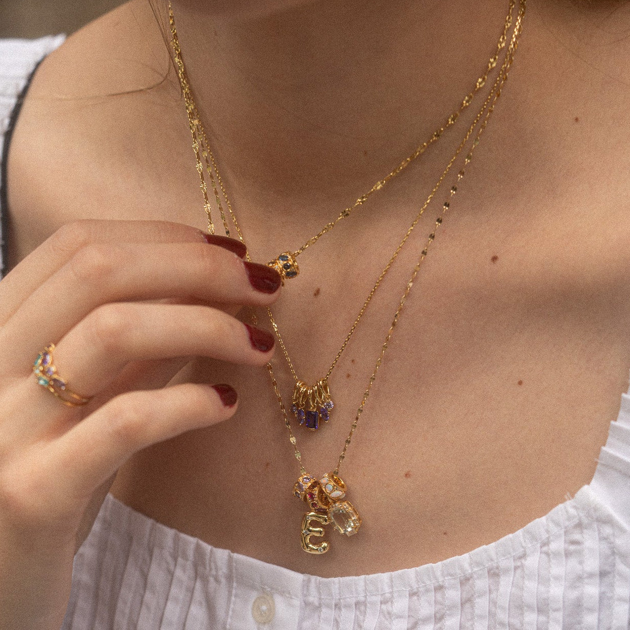 A woman in a white textured top showcases three layered gold necklaces, including the Star Crossed Celestial Bubble Letter Charm. She holds one necklace with red-manicured nails and wears a gold ring adorned with multicolored gemstones.
