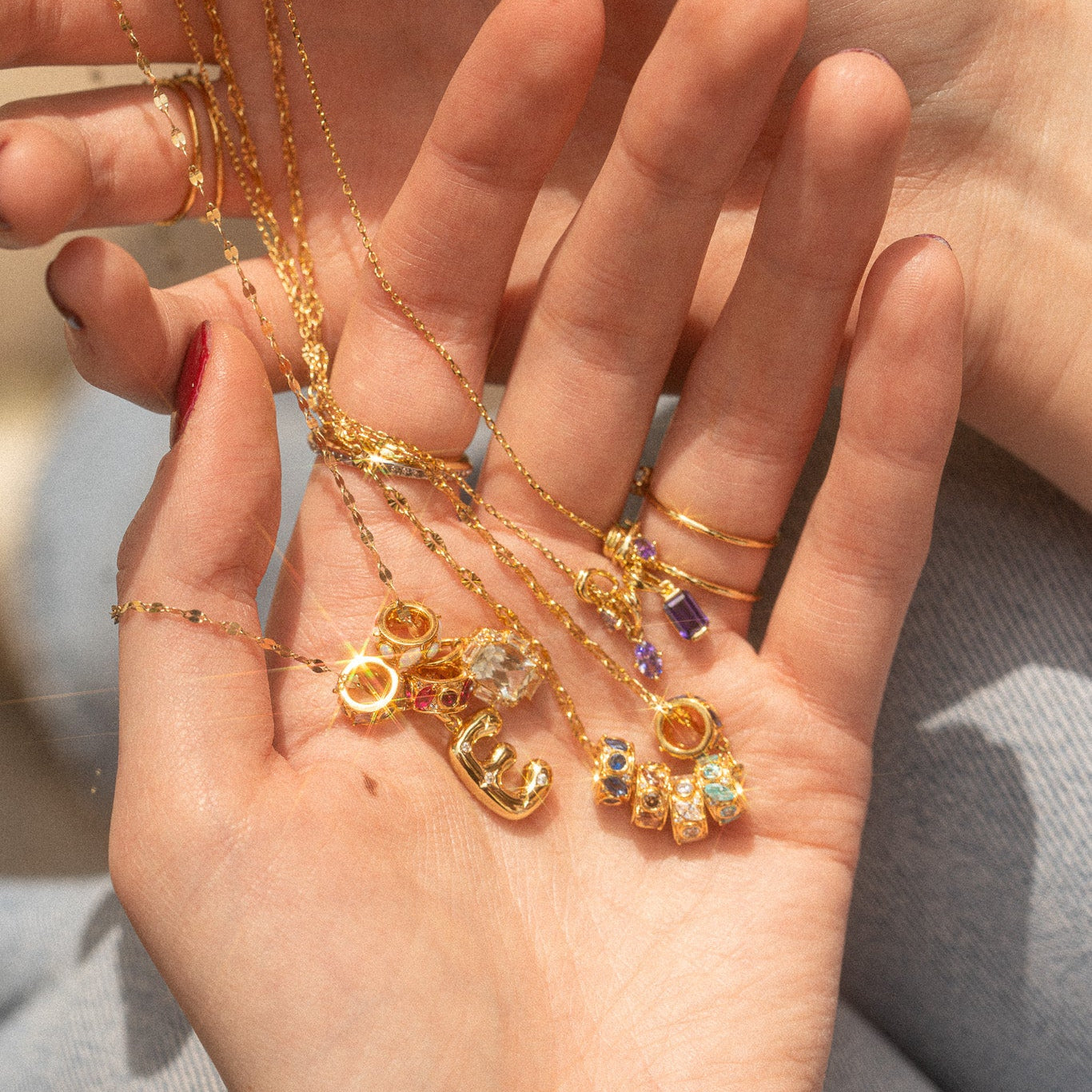A close-up of two hands holding gold necklaces and rings, featuring the Shades of You Birthstone Spacer Charm, with colorful gemstones catching the sunlight.