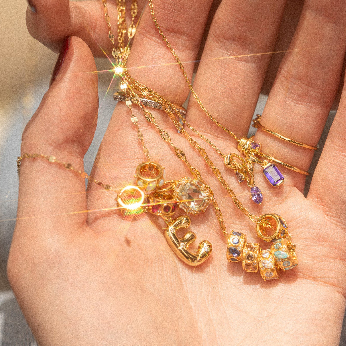 A close-up of a hand holding gold necklaces with gemstone pendants and dainty rings. Sunlight highlights the Star Crossed Celestial Bubble Letter Charm, making it shine among the sparkling jewelry.