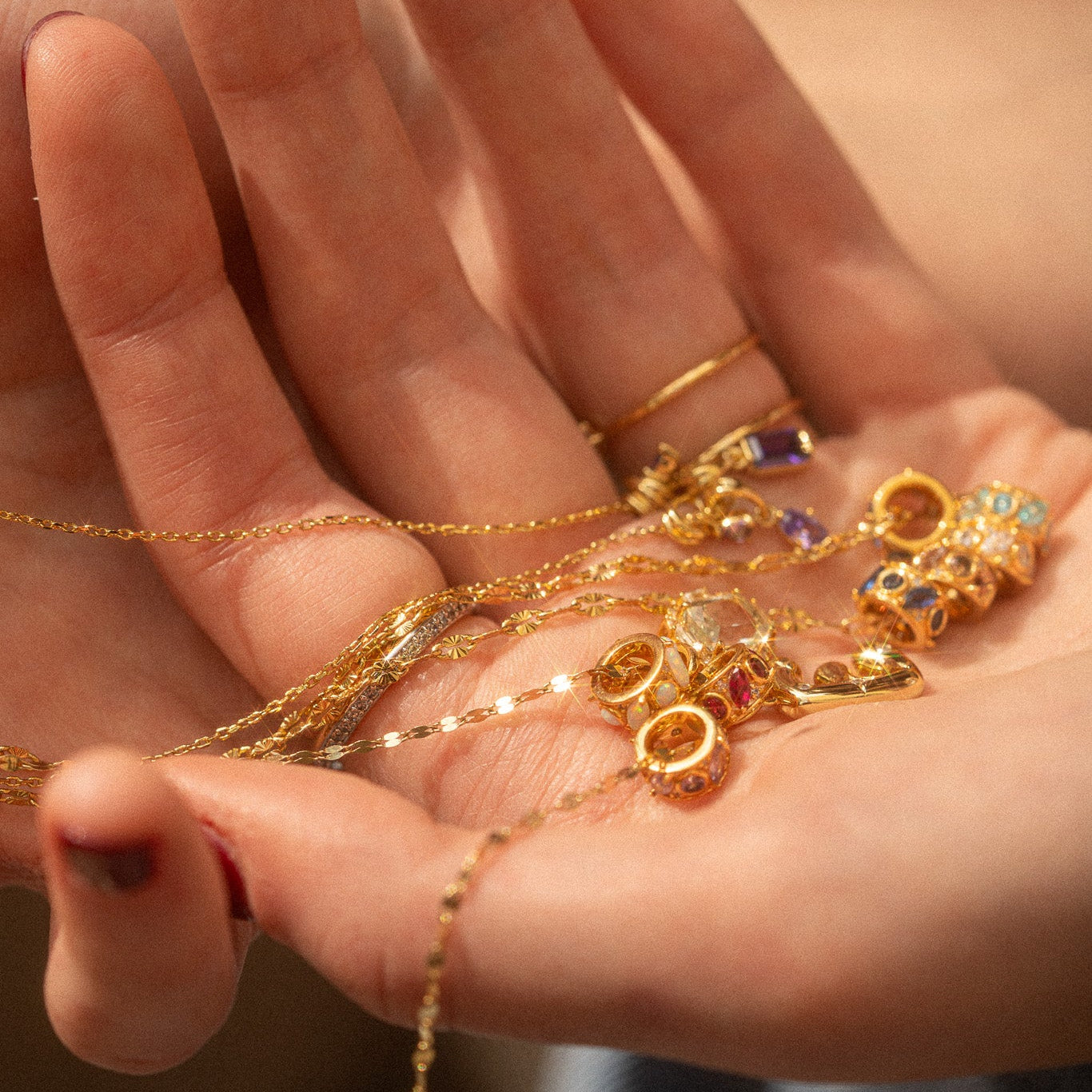 A hand holds several delicate gold necklaces with the "Shades of You Birthstone Spacer Charm" sparkling in natural sunlight; simple gold rings adorn the fingers.