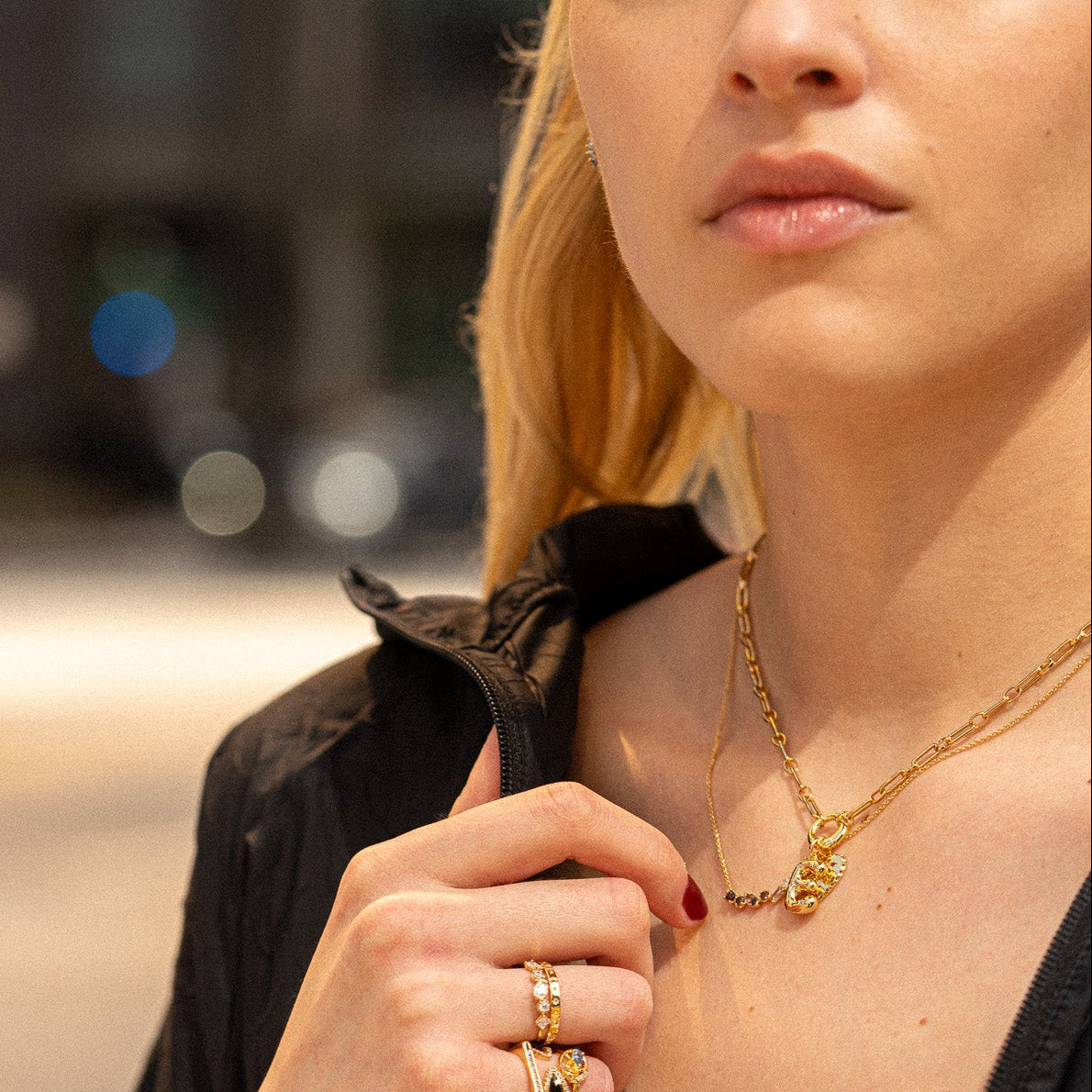 A woman with long blonde hair wears gold jewelry, including the Moon and Stars Stacking Ring, layered necklaces, and small earrings. She is dressed in a black jacket with light pink lipstick, set against a blurred background.