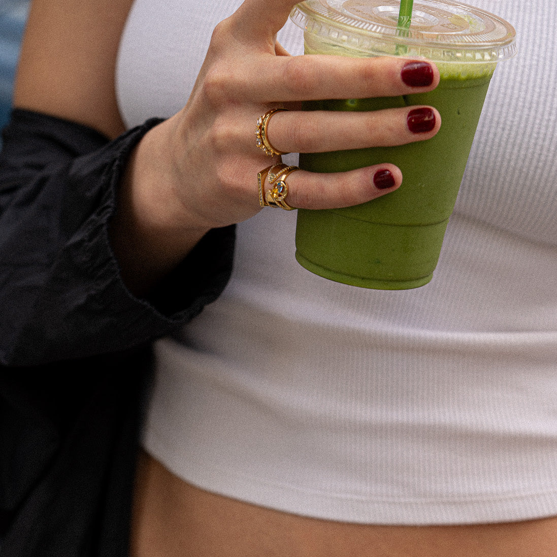 With maroon nails and an Eclectic Swirl Pinky Ring, a person holds a clear plastic cup of green smoothie while wearing a white ribbed tank top and a black sleeve draped over one arm.