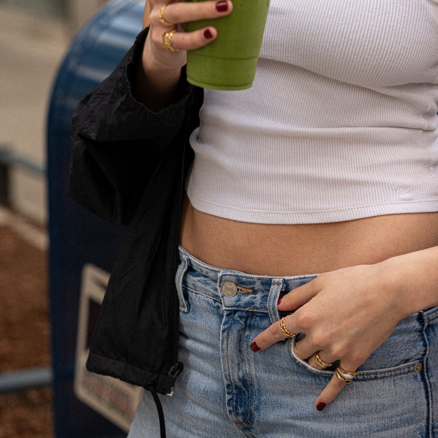 A person with red-painted nails wears multiple gold rings, including the Bold Toi Et Moi Pinky Ring, as they hold a green drink in one hand and touch their waist. They are dressed in a white crop top and blue jeans.