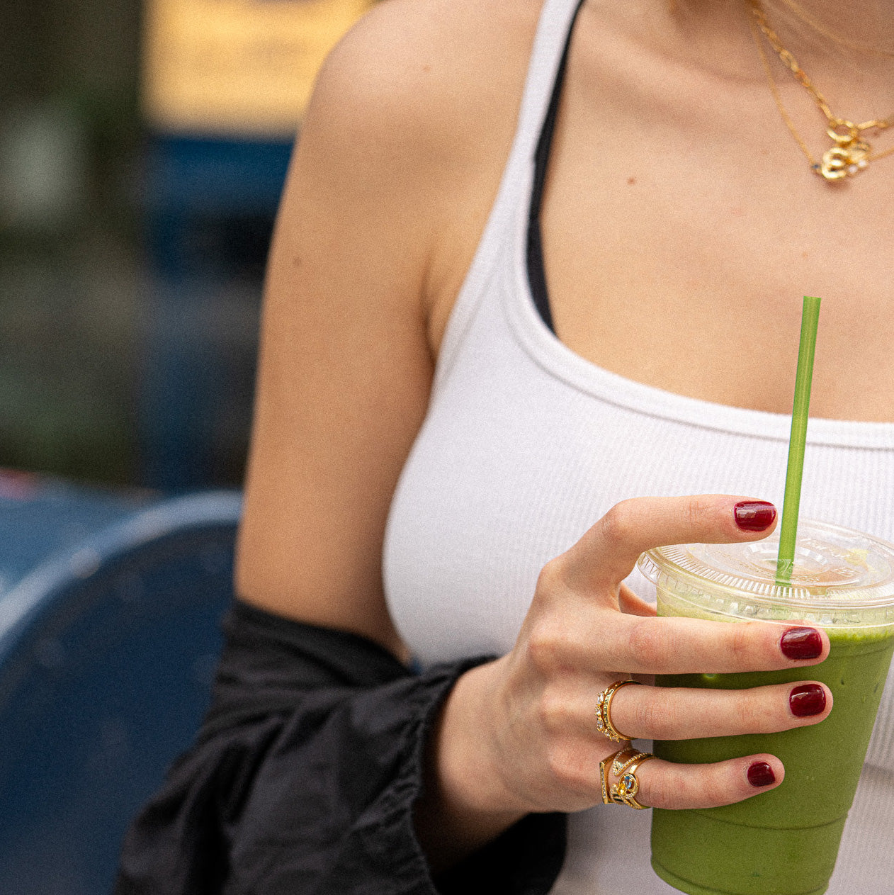 A woman wearing the Pave Bar Pinky Ring and layered necklaces holds a clear cup with a green smoothie and straw. She has red nails and is dressed in a white tank top.