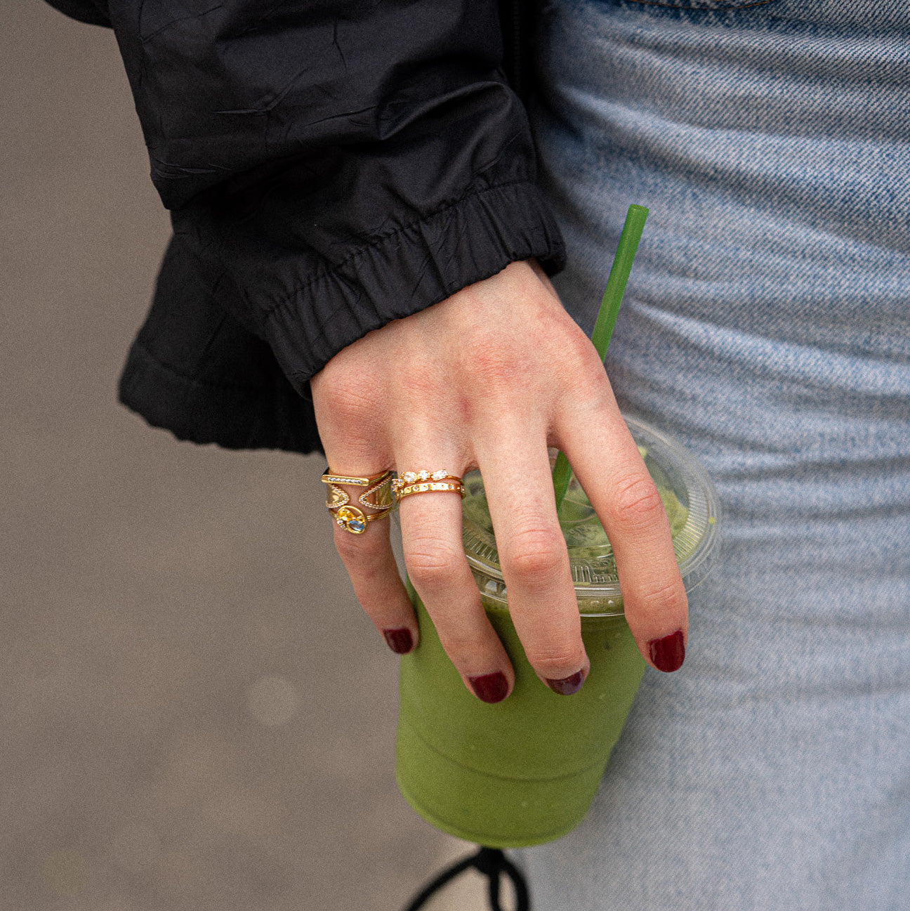 A person with red nail polish and an Open Front Cigar Pinky Ring holds a clear cup of green smoothie with a green straw, wearing a black jacket and light blue jeans.