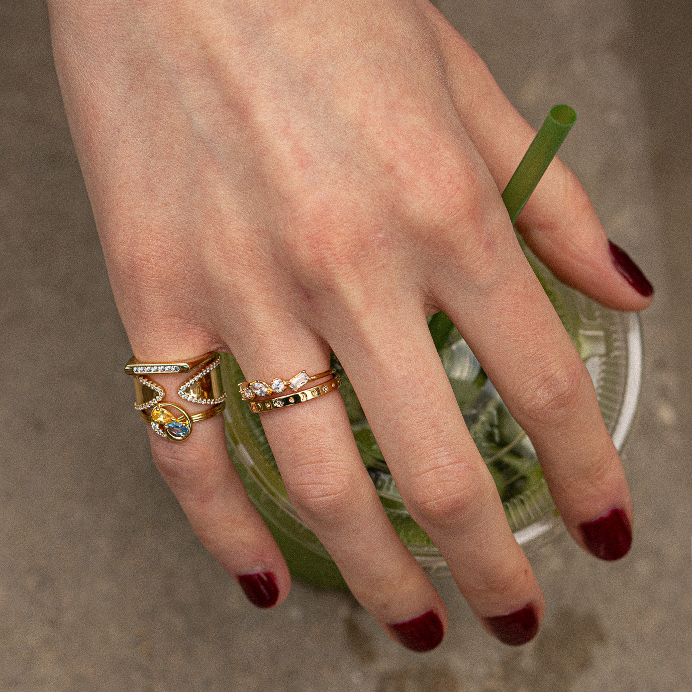 A hand with dark red nail polish wears the Eclectic Swirl Pinky Ring and holds a clear cup with a green straw, set against a blurred neutral background.