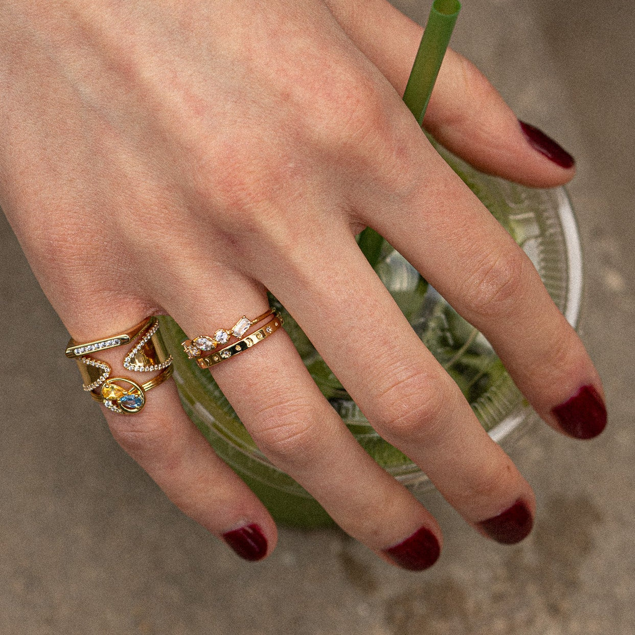 A hand with dark red nail polish holds a clear cup with a green drink and straw, wearing the Moon and Stars Stacking Ring.