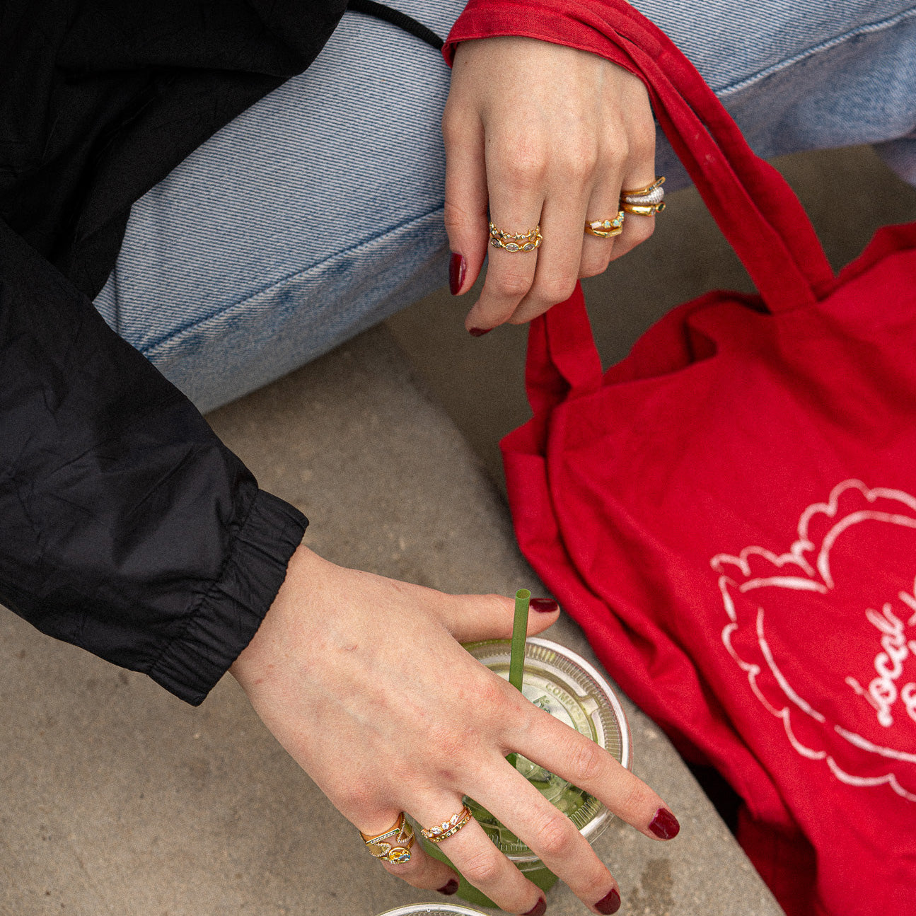 A person with red-painted nails wears the Pave Bar Pinky Ring and holds an iced drink with a green straw. They are dressed in blue jeans, a black top, and have a red tote bag featuring a white floral design on their lap.