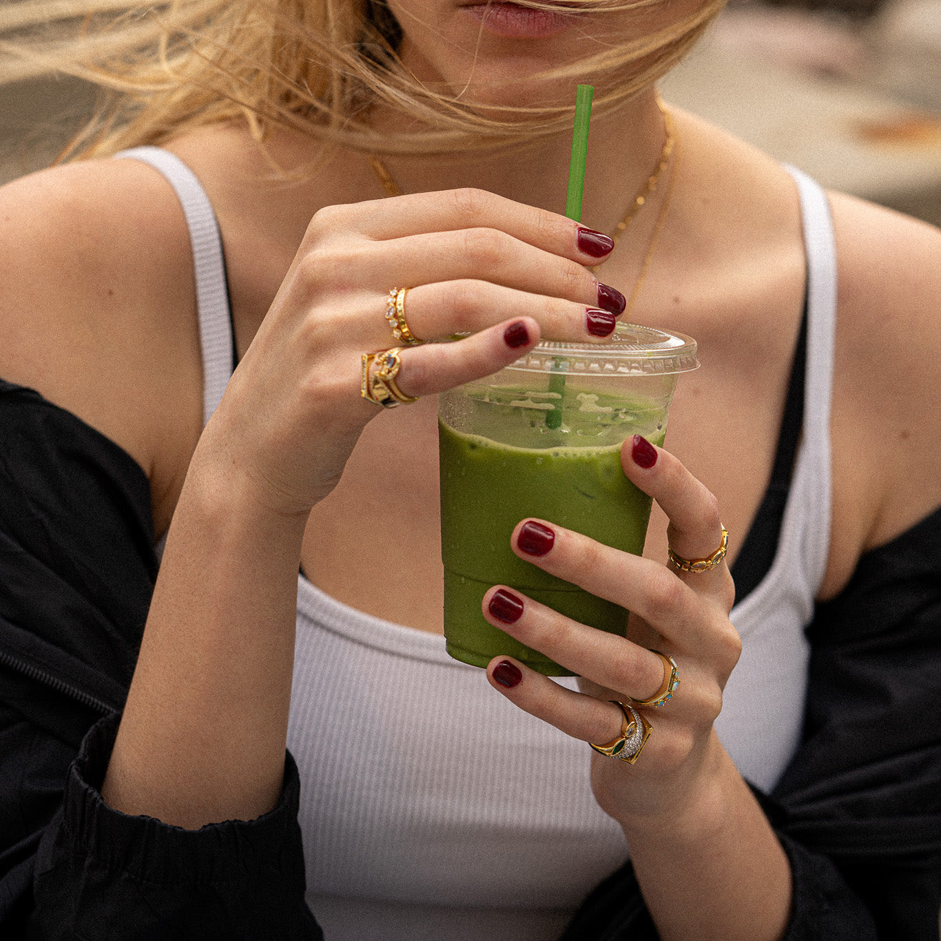 A woman with painted red nails wears the Bold Toi Et Moi Pinky Ring as she holds a plastic cup of green smoothie with a green straw. She is dressed in a white tank top and has a black jacket draped off her shoulders, her face partially out of frame.