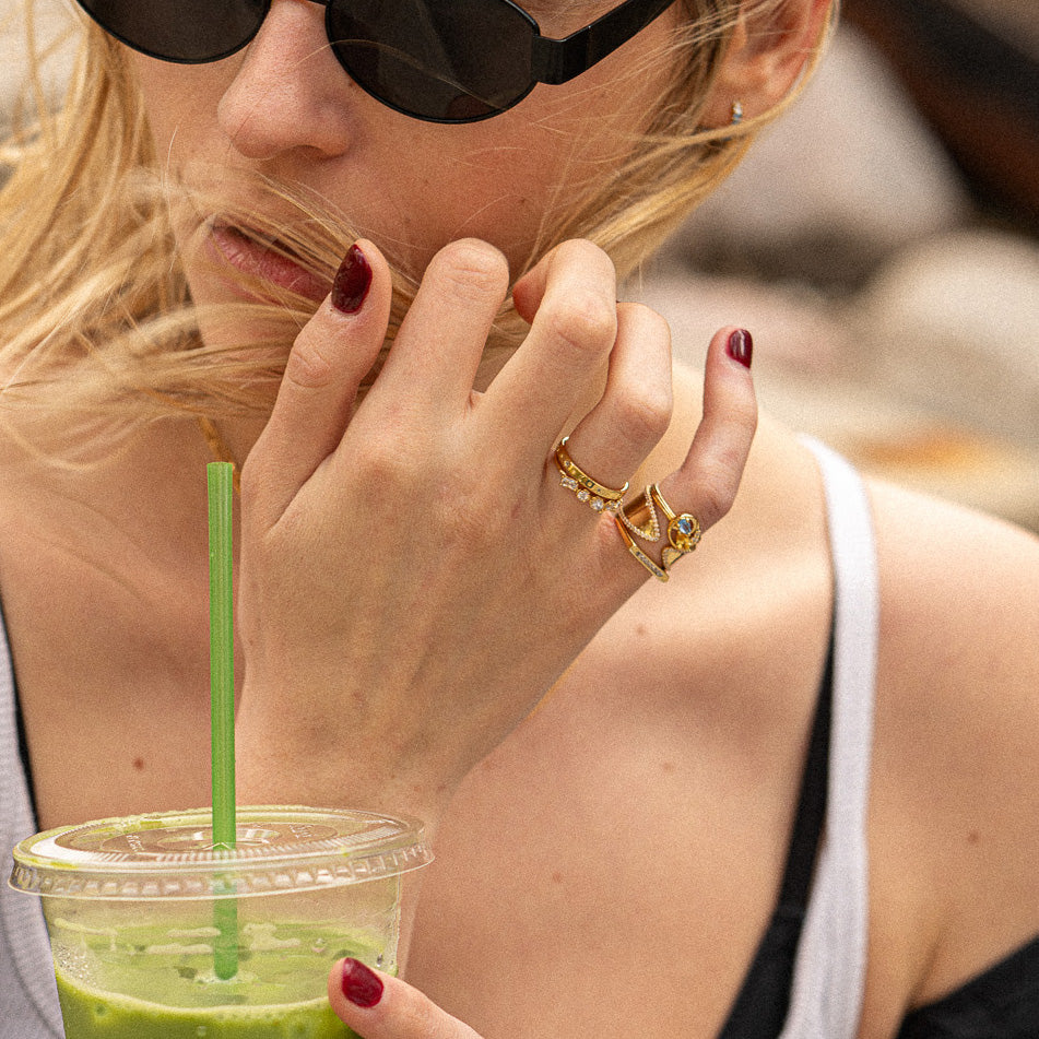 A woman with windswept hair and red nails wears the Eclectic Swirl Pinky Ring and sunglasses as she holds a green smoothie with a straw.