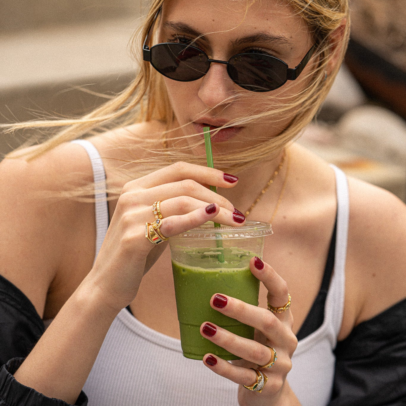 A woman with red nail polish and blond hair partly covering her face sips a green smoothie, styling black sunglasses, gold necklaces, and the Eclectic Swirl Pinky Ring among other gold rings.