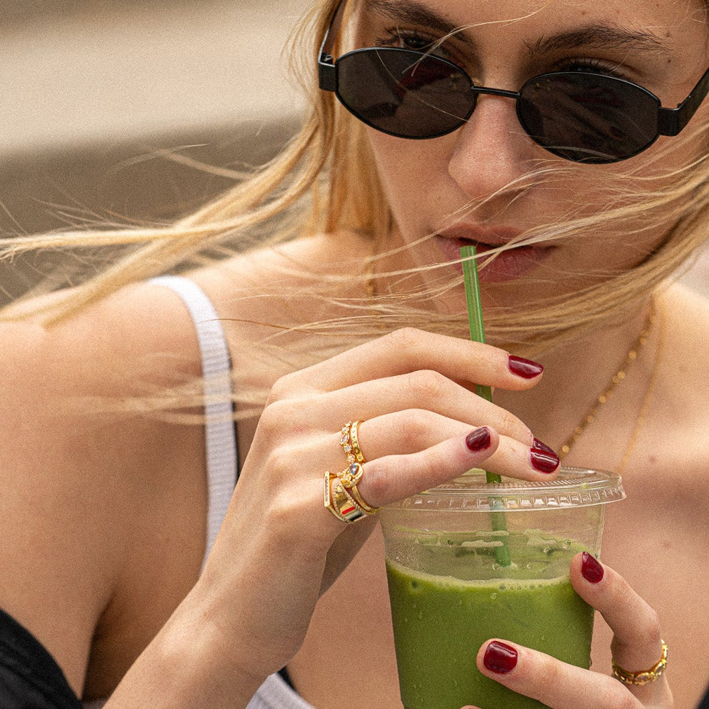 A woman with blonde hair in a white tank top and red nail polish sips a green smoothie, wearing dark sunglasses, gold jewelry, and the Moon and Stars Stacking Ring.