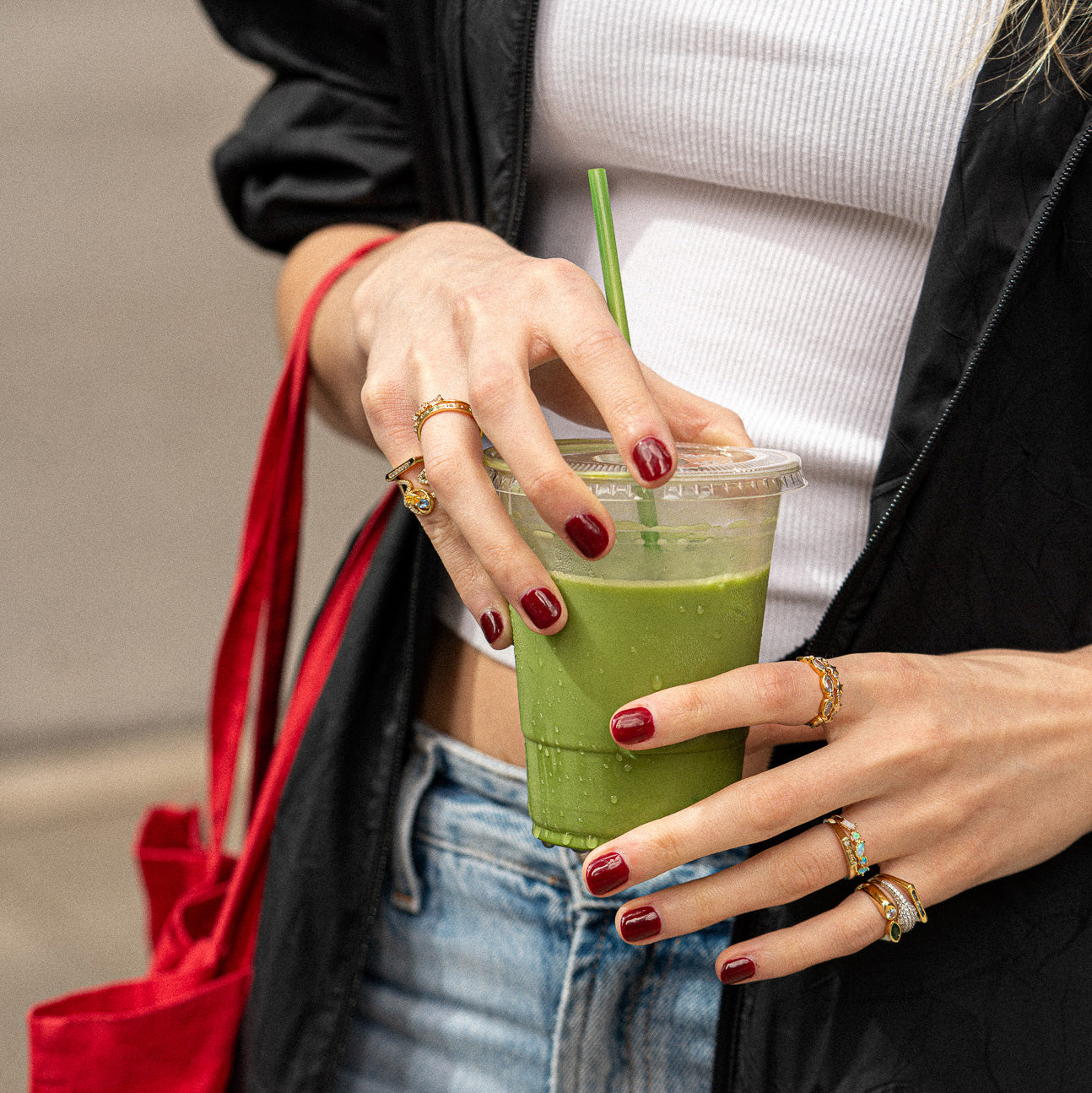 Wearing a white tank top, denim shorts, and a black jacket, the person holds a green smoothie and carries a red bag. They wear red nail polish and accessorize with gold rings, including the Pave Signet Pinky Ring.