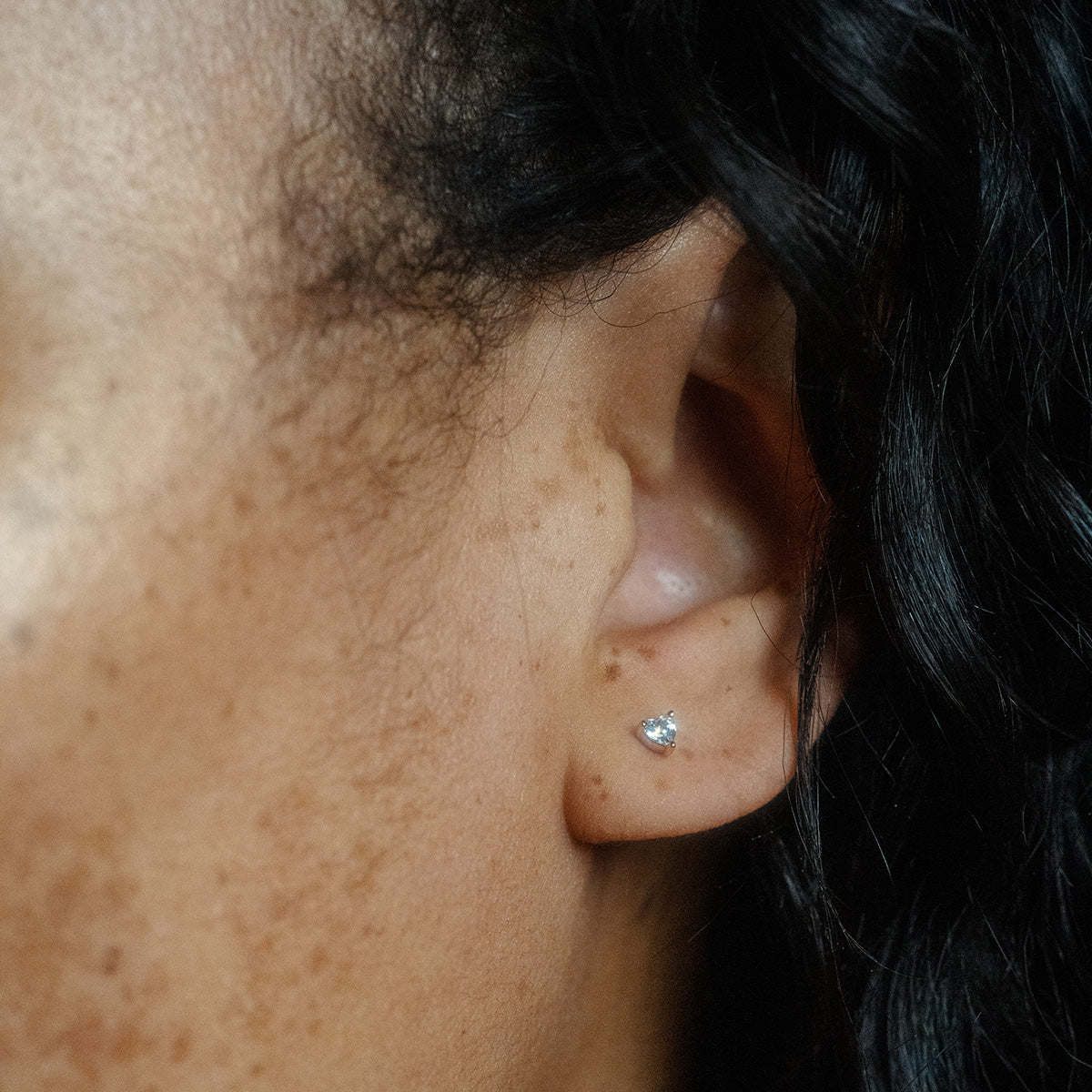 A close-up of a person’s ear with a silver stud earring and curly hair—perfect inspiration for gifting the Limited Edition Valentine’s Day Solid Gold Hearts Gift Set.
