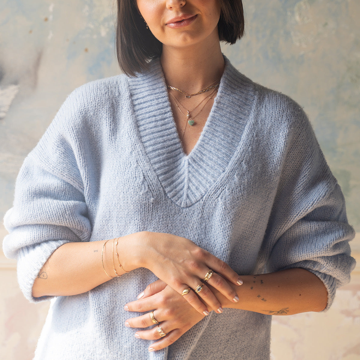 A woman in a light blue, oversized knit sweater with a deep V-neck wears gold necklaces, bracelets, and the Solid Gold Aurora Angel Aura Quartz and Blue Topaz Ring, standing with arms crossed and smiling softly in natural light.