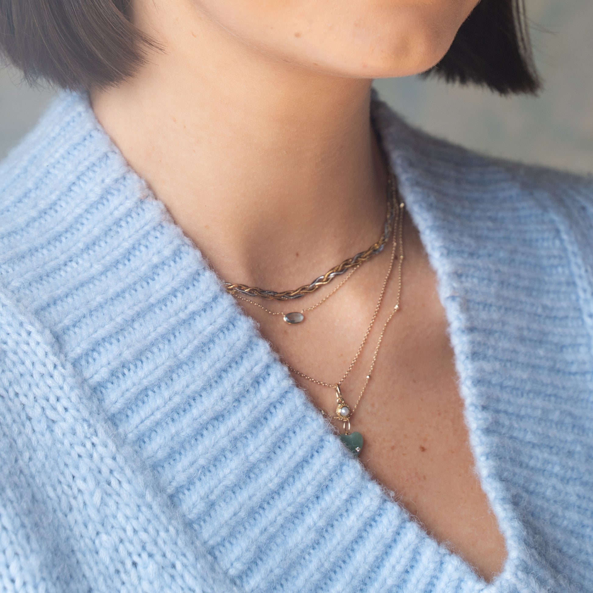 A person wears a light blue knit sweater with a deep V-neckline, showing layered gold necklaces—one is the Solid Gold Daybreak Swiss Blue Topaz Necklace. Only the lower face, neck, and upper chest are visible.