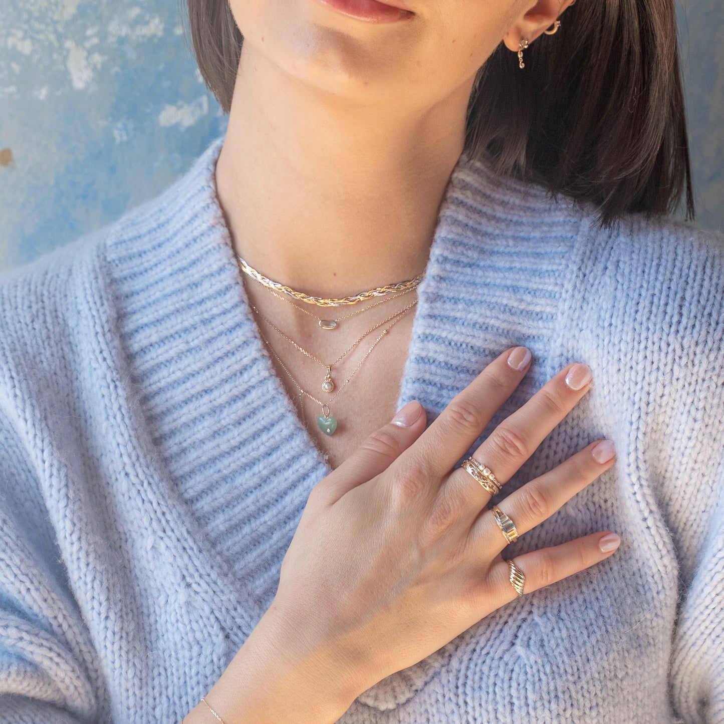 A woman in a light blue knit sweater showcases layered Family Gold necklaces, featuring the Solid Gold Daybreak Swiss Blue Topaz Necklace, along with gold rings and small hoop earrings. Her hand rests on her chest, highlighting the elegant jewelry.