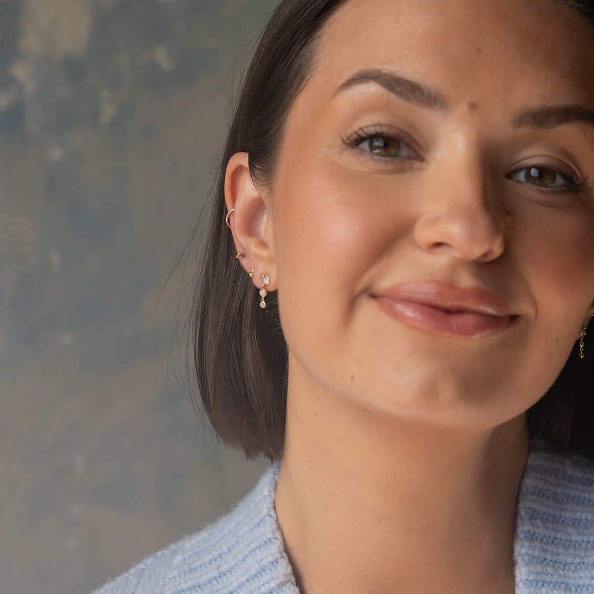 A woman with straight dark hair and natural makeup smiles softly, wearing Family Gold Solid Gold Diamond Dawn Ball Studs. She is dressed in a light blue sweater, set against a neutral, softly blurred background.