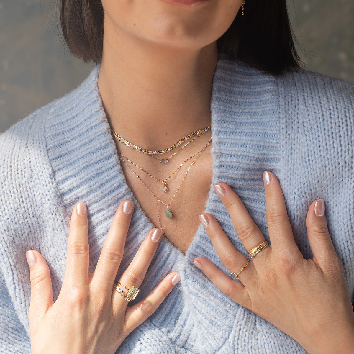 A woman in a light blue knit sweater showcases layered Family Gold necklaces, several gold bands, and the Solid Gold First Light Pastel Gemstone Ring, highlighting her manicured nails painted with light pink polish.