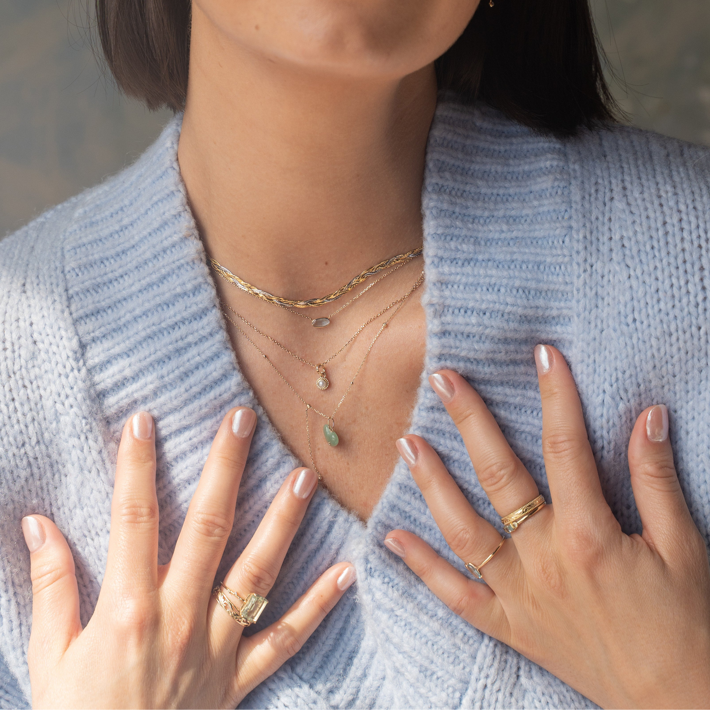 A woman in a light blue knit sweater wears the Solid Gold Dewdrop Green Amethyst Statement Ring, paired with layered gold necklaces featuring small pendants. Her naturally manicured hands, adorned with elegant jewelry, rest gracefully near her collar.