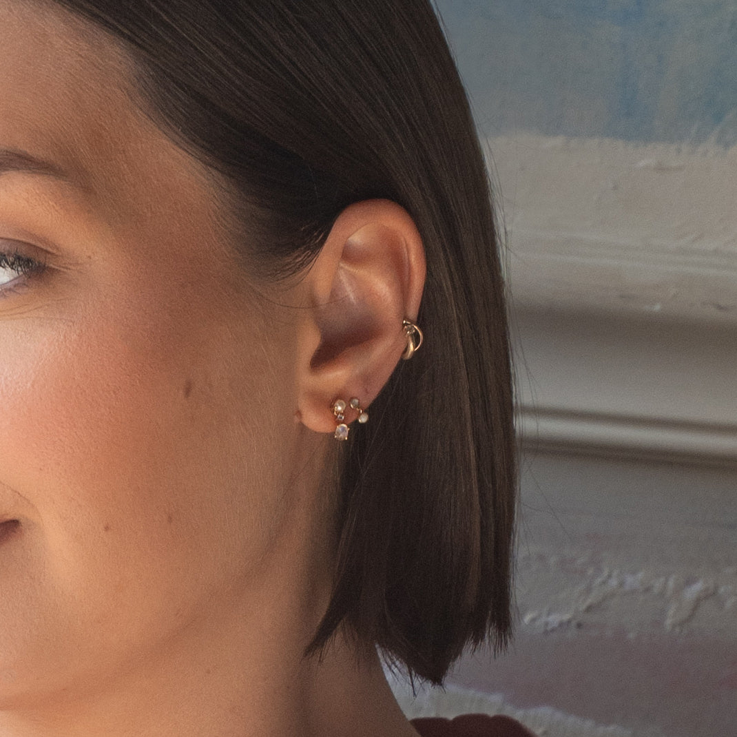 Close-up of a woman's ear adorned with multiple piercings, including Solid Gold Soft Glow Pearl and Moonstone Drop Studs. Brown hair is tucked back, revealing her face and neck against a softly textured background.