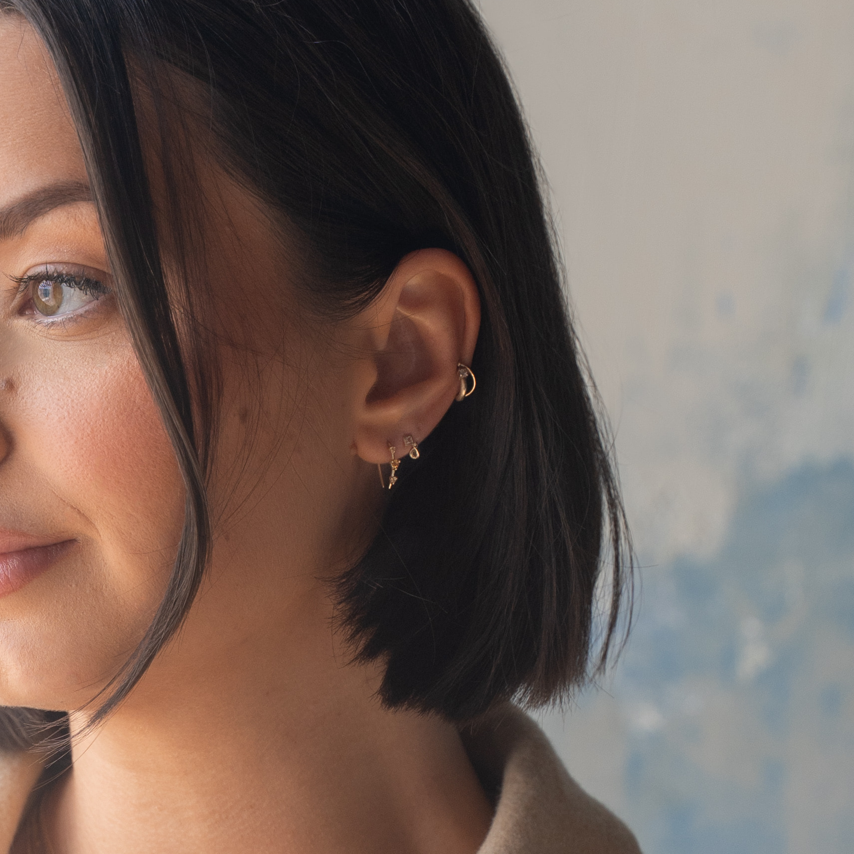 A woman with straight dark hair and gold hoop earrings faces left in close-up, showing clear skin, natural makeup, and Solid Gold Radiant White Topaz Drop Studs in her ear stack. The background is softly blurred in neutral tones.