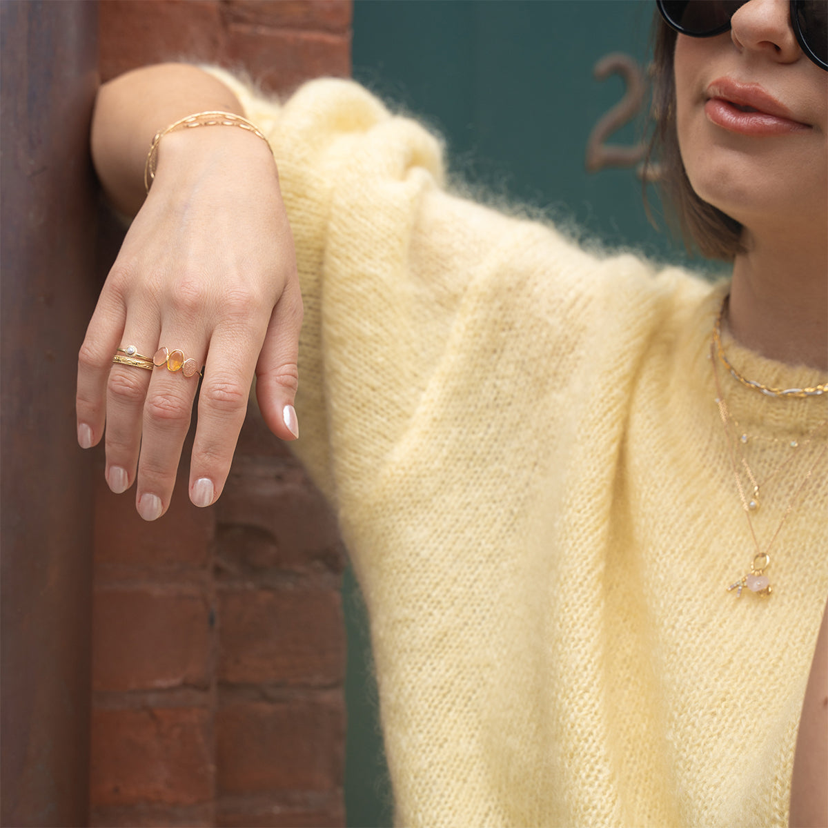 A woman in a fuzzy yellow sweater leans against a brick wall, wearing sunglasses and layered gold jewelry, including the Solid Gold Morning Light Citrine and Sunstone Ring, along with bracelets, necklaces, and a chain. Her nails are neutrally manicured.
