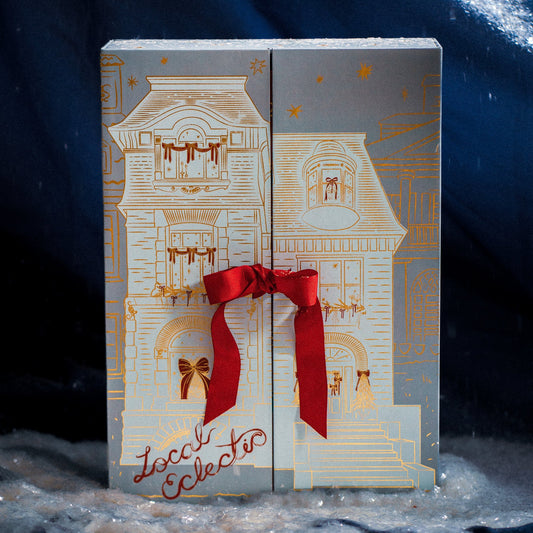 A decorative box with gold house and window designs, tied with a red ribbon, rests on snow. “2025 Jewelry Advent Calendar” appears in red script at the bottom left.