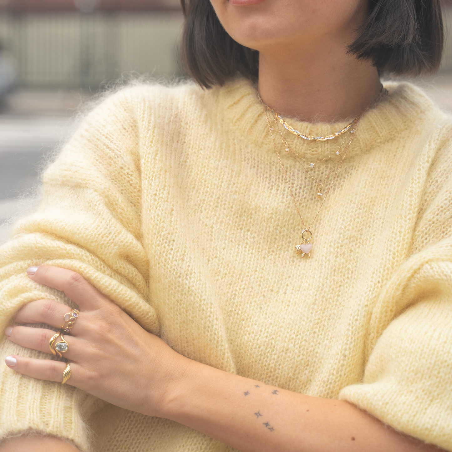 A person wears a fuzzy yellow sweater, gold rings, bracelets, and the Solid Gold Horizon Moonstone and Opal Sprinkle Necklace. Small star tattoos are visible on their forearm. The image focuses on their torso and crossed arms; face partially out of frame.