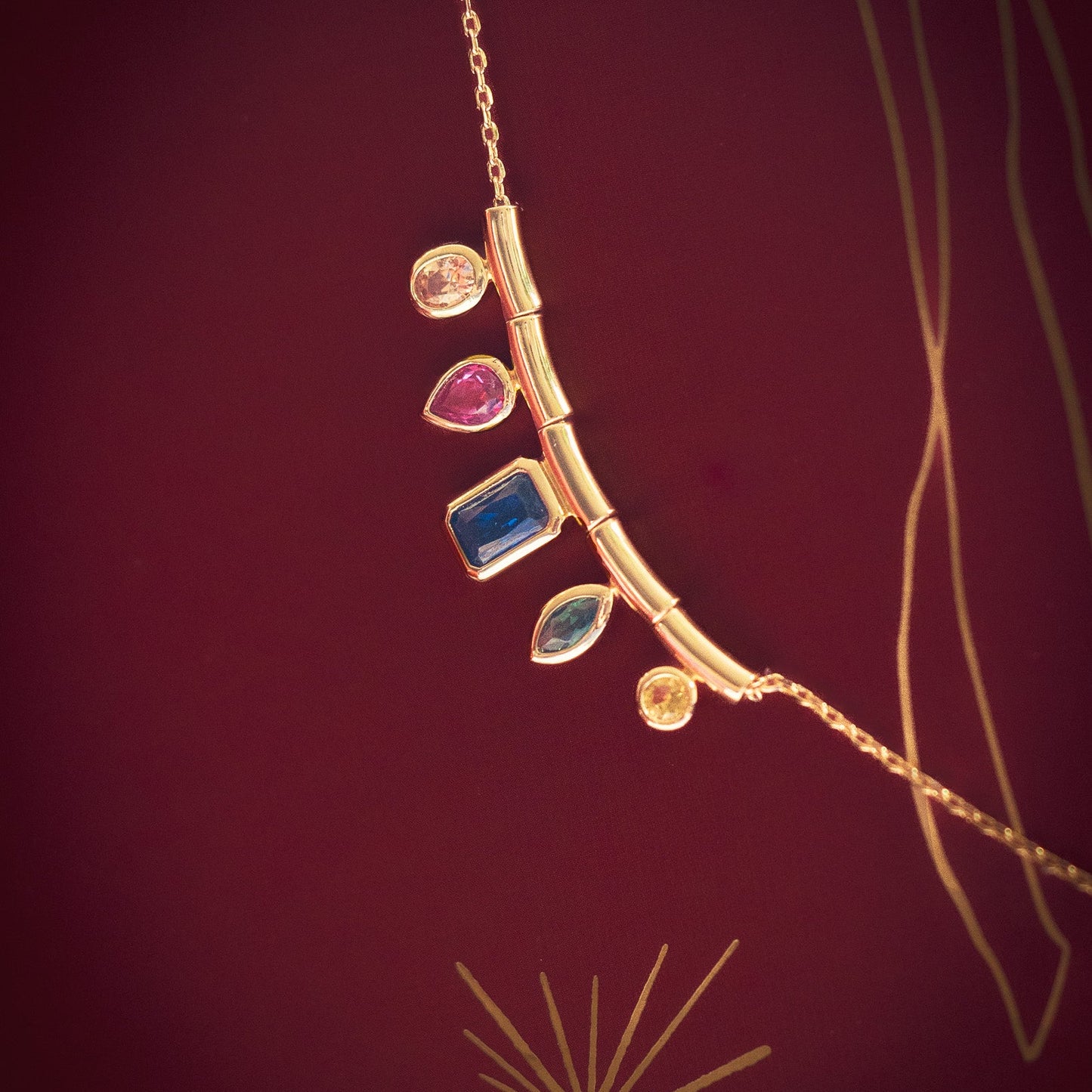 The Technicolor Gem Drop Necklace is a gold piece with a curved bar pendant showcasing five uniquely shaped and colored gemstones, displayed on a deep red background with elegant gold line accents.