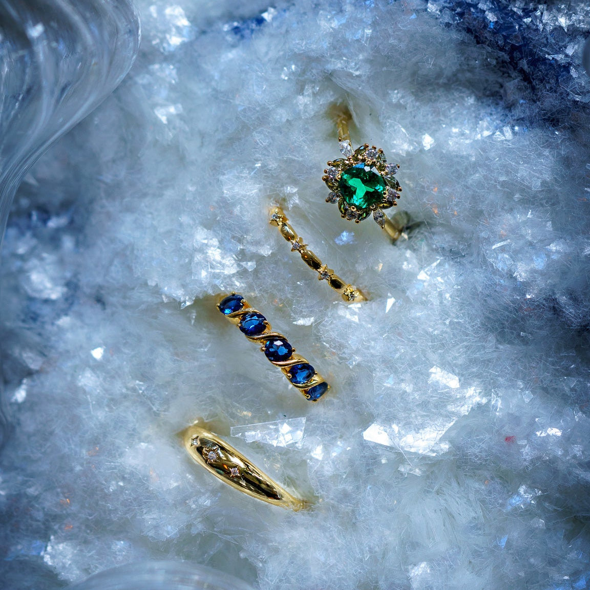 Three gold rings with blue and green gemstones shine on a sparkling surface, capturing the magic of the 2025 Jewelry Advent Calendar—perfect holiday gifts for jewelry lovers.
