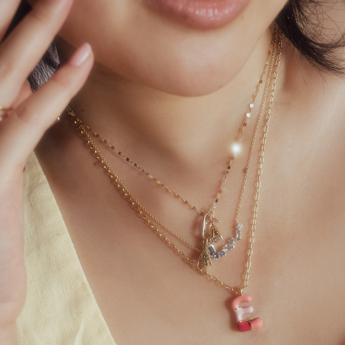 A close-up of a person with light skin wearing a light top and the Shades of You Ombre Birthstone Necklace, layered with gold pendants including a pink and white beaded letter S.