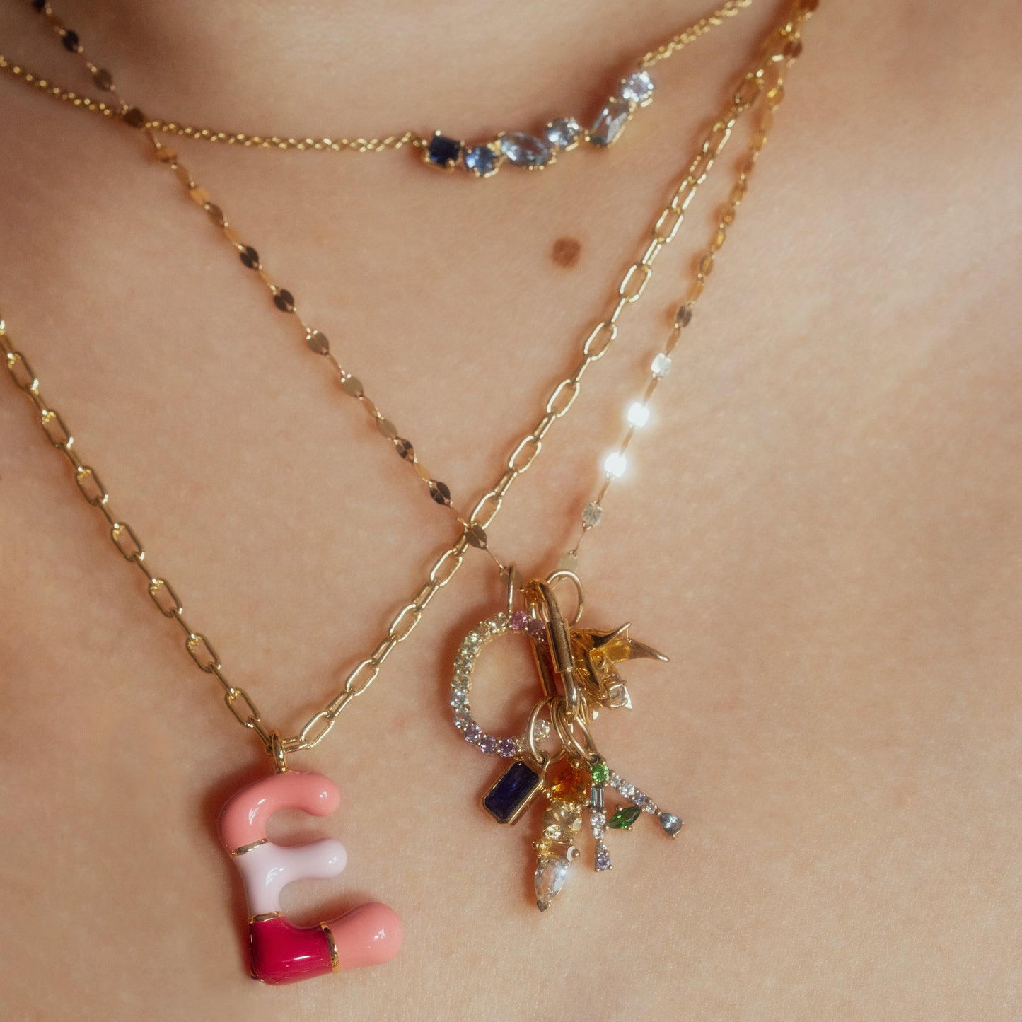 Close-up of layered gold necklaces on bare skin, featuring the Solid Gold Pastel Gemstone Letter Charm with pink and red accents, alongside another necklace adorned with pastel gemstone charms spelling CORK.