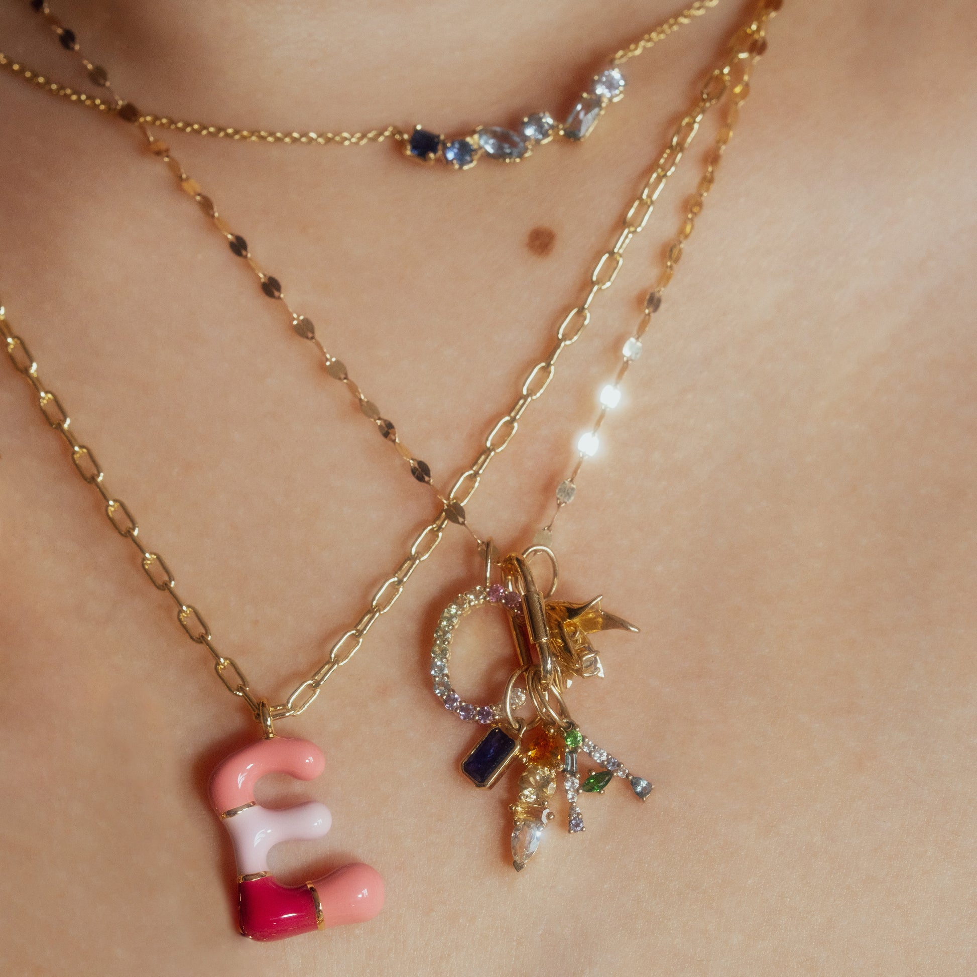 Close-up of layered gold necklaces on bare skin, featuring the Solid Gold Pastel Gemstone Letter Charm with pink and red accents, alongside another necklace adorned with pastel gemstone charms spelling CORK.