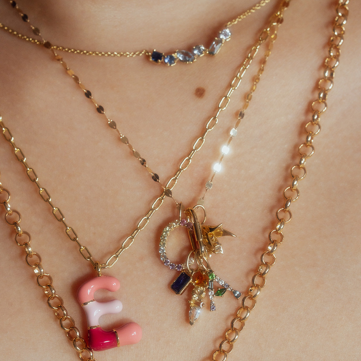 A close-up of a neck adorned with gold chain necklaces, featuring colorful pendants like a pink and red letter E, and the Solid Gold Birthstone Baguette Charm.