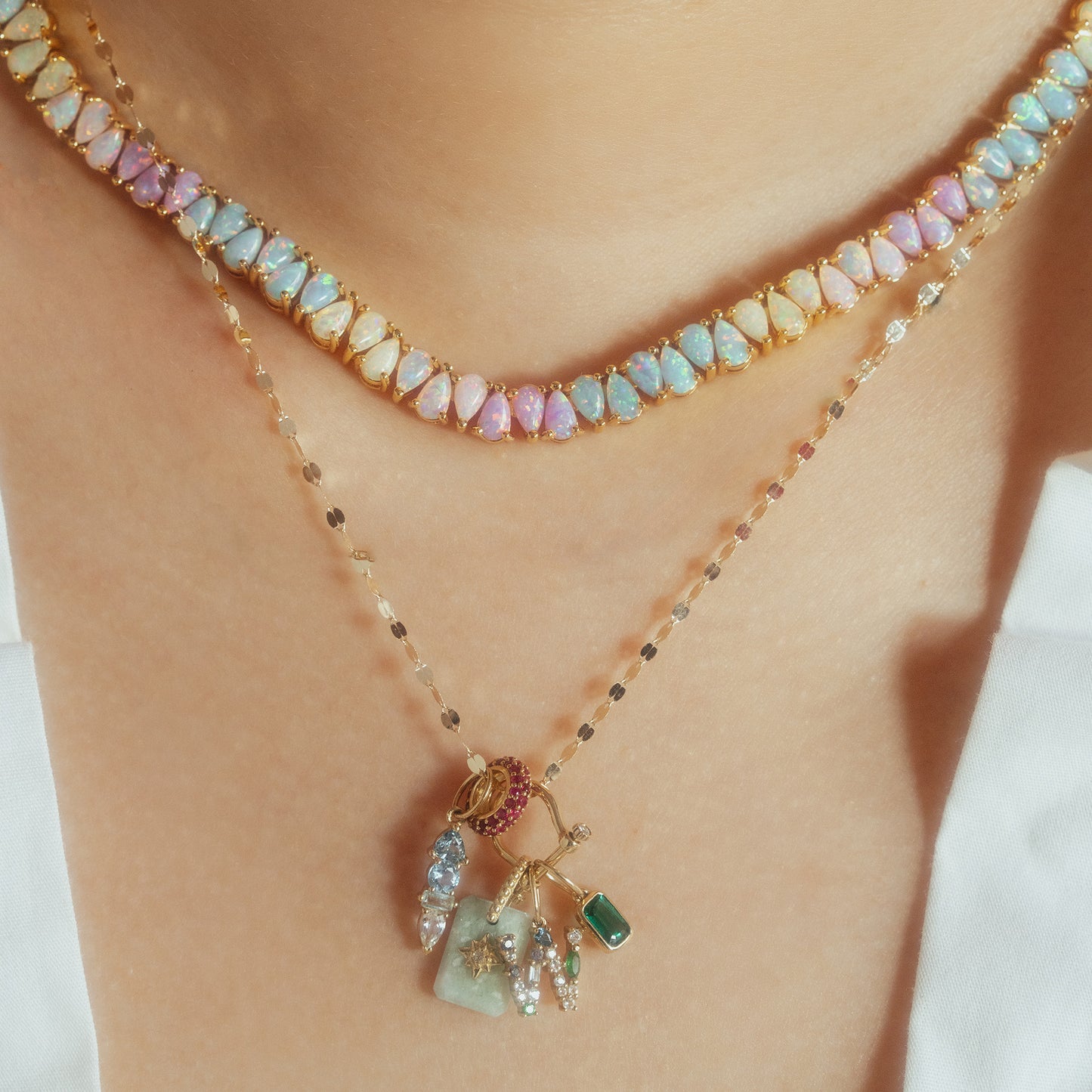 A close-up of two layered necklaces: one with pastel opal-like stones set in gold, and the other, the Solid Gold Modern Ombre Birthstone Charm, featuring a gold chain with colorful gemstone charms, worn on light skin.